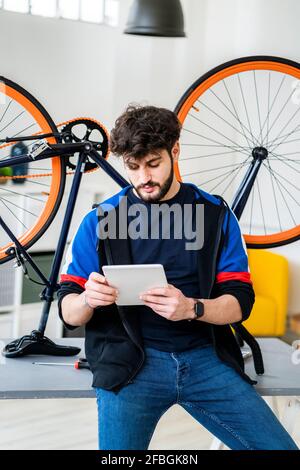 Man using digital tablet while leaning on table with upturnt bicycle at home Stock Photo