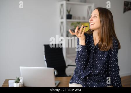 Smiling female employee is using voice assistant on the smartphone, young woman talking in the mic of her mobile phone, recording voice message, using voice recognizable function or app Stock Photo