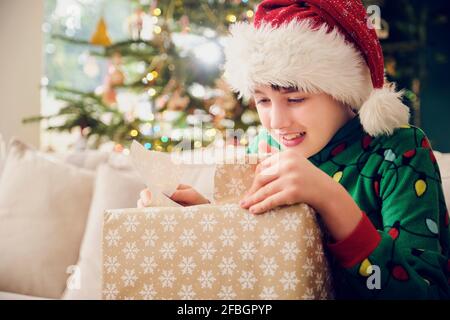 Cute boy in Santa hat unwrapping gift during Christmas at home Stock Photo