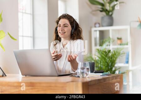 Smiling female woman gesturing while talking through headphones at home office Stock Photo
