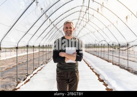 Smiling farmer with arms crossed holding digital tablet in agricultural field Stock Photo