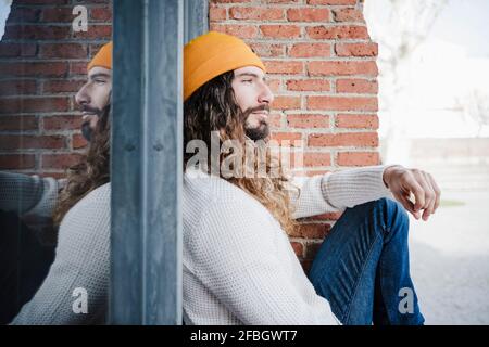 Man wearing knit hat sitting by brick wall while looking away Stock Photo