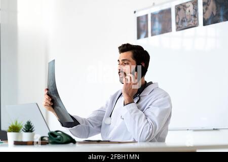 Male healthcare worker examining X-ray while talking on smart phone at hospital Stock Photo