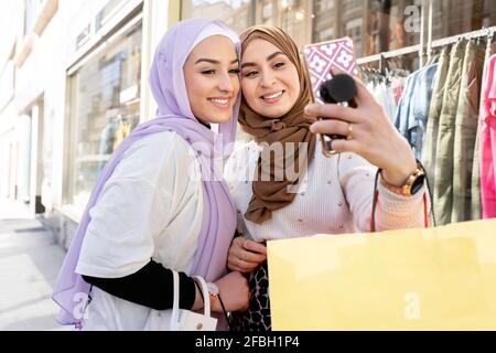 Female Arab friends in hijabs taking selfie through smart phone while shopping in city Stock Photo