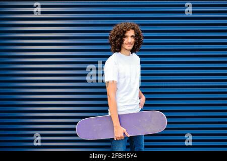 Curly haired man holding skateboard while standing in front of blue shutter Stock Photo