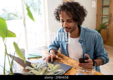 Young businessman holding smart phone while using laptop at home Stock Photo