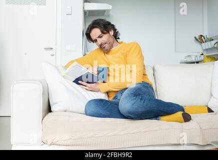 Man reading book while sitting on sofa in living room at home Stock Photo