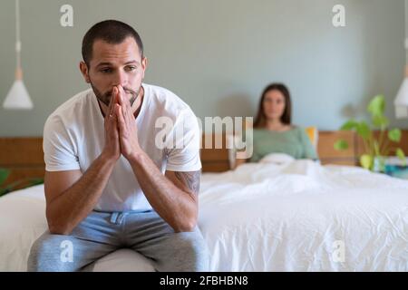 Serious man sitting on bed with girlfriend in background at home Stock Photo