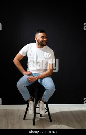 Smiling man looking away while sitting on stool by black wall Stock Photo
