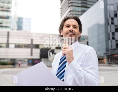 Smiling male architect holding blueprint while standing in office park Stock Photo