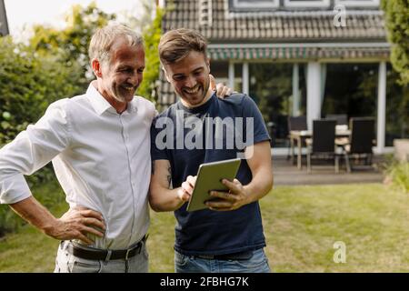 Happy son using digital tablet while standing by father in backyard Stock Photo