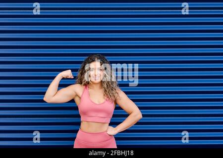 Confident young female sportsperson flexing muscle in front of blue wall Stock Photo