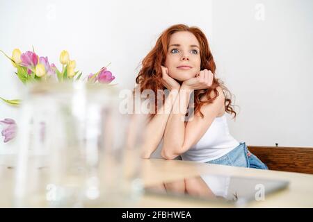 Thoughtful redhead mid adult woman sitting with hands on chin while leaning on table at home Stock Photo