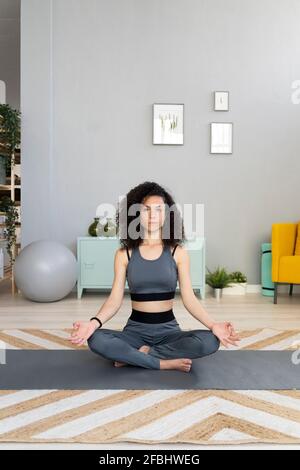 Young woman practicing lotus position on exercise mat in living room