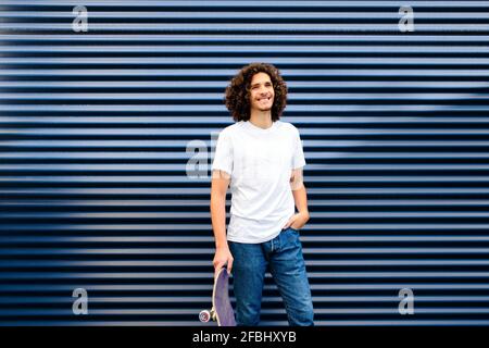 Young man with curly hair holding skateboard while standing in front of blue shutter Stock Photo