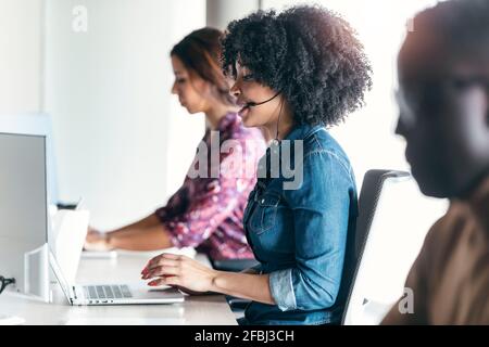 Female customer service representative using laptop while sitting amidst colleagues at office Stock Photo