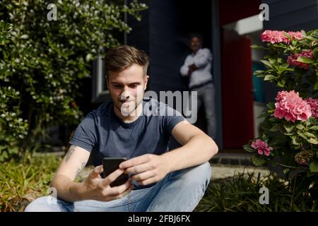 Son using smart phone while father leaning on wall Stock Photo