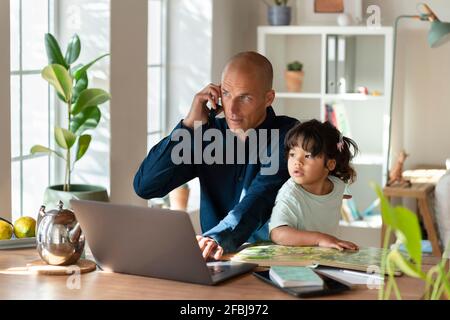 Father talking on mobile phone while sitting with daughter in home office Stock Photo