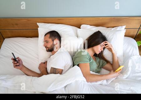 Young couple using smart phones while lying back to back in bed at home Stock Photo
