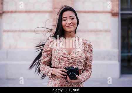 Smiling woman with eyes closed holding camera by wall Stock Photo