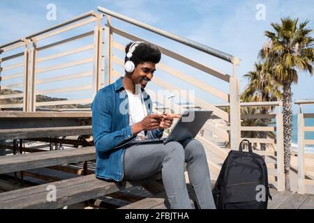 Happy man gesturing during video call on laptop while sitting on promenade during sunny day Stock Photo