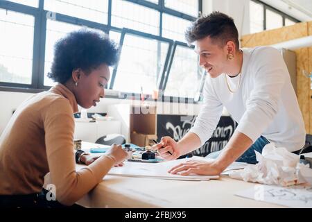 Smiling hipster man teaching girlfriend calligraphy while sitting at table in studio Stock Photo
