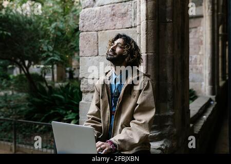 Contemplating man with long hair looking up while sitting with laptop in front of wall Stock Photo
