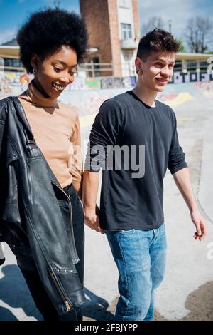 Smiling multi ethnic couple walking while holding hands at skateboard park during sunny day Stock Photo
