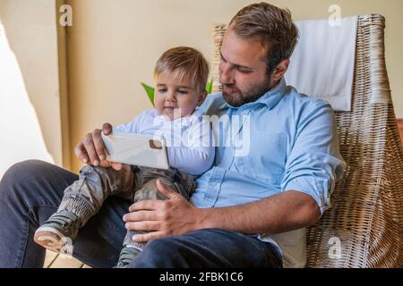 Father sharing mobile phone with cute son sitting on lap at home Stock Photo