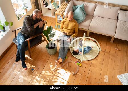 Two men looking at playing baby girl on floor in living room Stock Photo