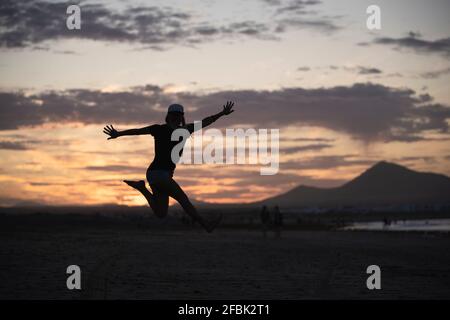 Excited woman with arms outstretched jumping at beach during sunset Stock Photo