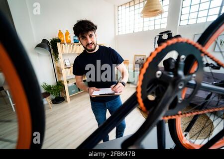 Smiling man with clipboard repairing bicycle at home Stock Photo