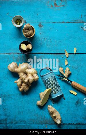 Ginger root, grater, small cups and kitchen knife lying on blue wooden surface Stock Photo