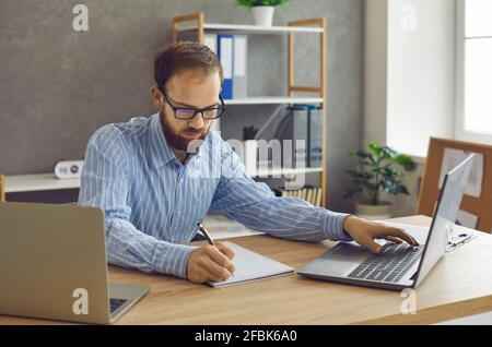 Serious concentrated man sitting at desk with laptops and taking notes in notebook Stock Photo