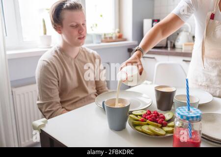 Woman pouring milk in cup for teenage son at dining table in kitchen Stock Photo