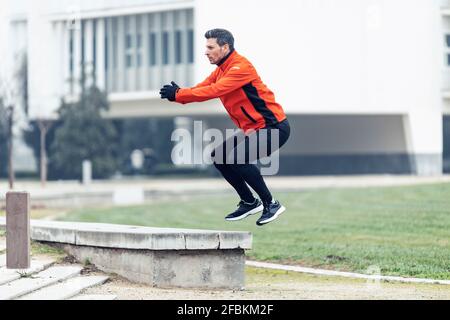 Sporty man doing jumping squats on retaining wall at public park Stock Photo