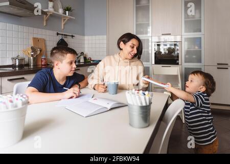 Boy giving book to mother while brother doing homework at table in kitchen Stock Photo