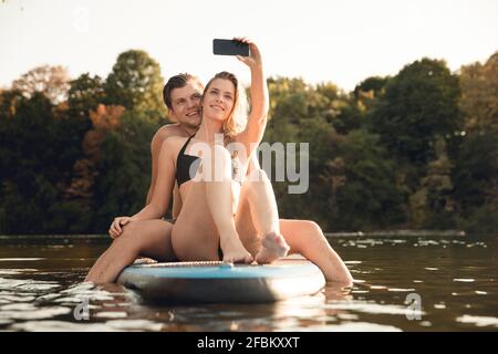 Happy couple sitting on paddleboard on a lake, taking smartphone selfies Stock Photo