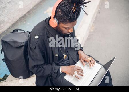 Young freelance worker with headphones using laptop while sitting on footpath Stock Photo