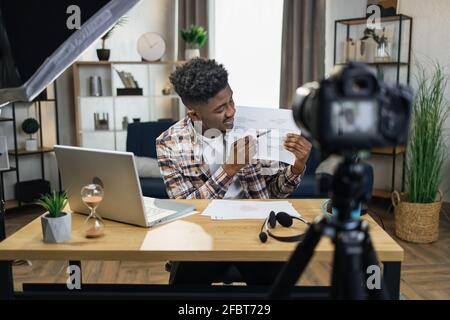 Focused african tutor recording masterclass for students using video camera at home. Young man in casual wear showing and explaining various graphs. Stock Photo