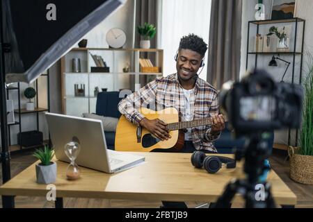 Happy african guy in headset sitting at table and recording video tutorial how to play guitar. Male musician creating online lessons while staying at home. Stock Photo