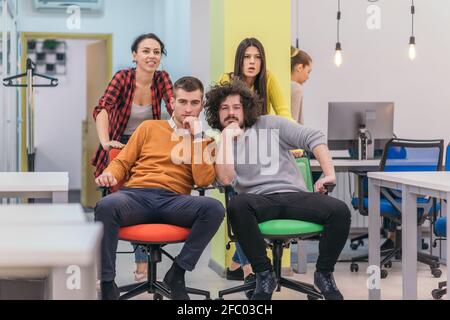 Team building and office fun.Four young cheerful businesspeople in smart casual wear having fun while racing on office chairs and smiling. Stock Photo