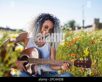 Cheerful woman with curly white hair and tattoos playing the guitar in a flower field Stock Photo