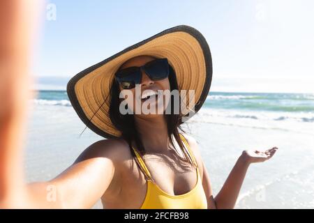 Smiling mixed race woman wearing sunhat and sunglasses taking selfie on beach Stock Photo