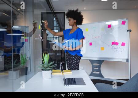 Mixed race businesswoman sticking memo notes on glass wall with whiteboard in background Stock Photo