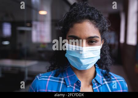 Portrait of mixed race businesswoman wearing face mask standing in corridor in office Stock Photo
