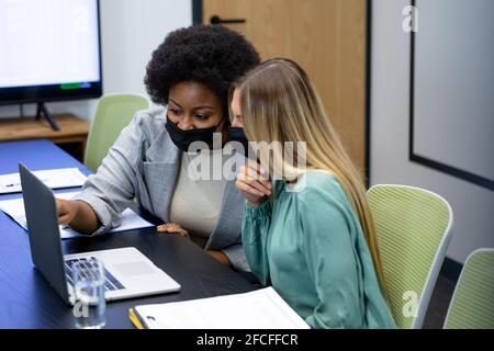 Diverse female business colleagues wearing masks talking and using laptop in meeting room Stock Photo