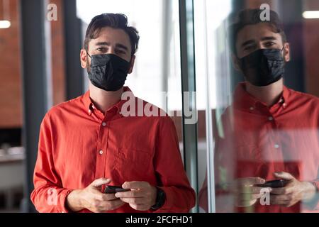 Portrait of caucasian businessman wearing face mask standing in corridor using smartphone Stock Photo