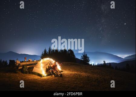 Couple of travelers set up camp on night mountain lawn and relaxing in iluminated tourist tent after dinner. Light of surrounding villages, mountain peaks under evening starry sky on the background. Stock Photo