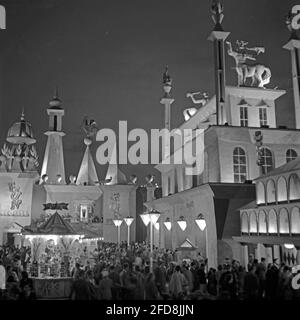 Evening crowds mingle at the Piazza, Battersea Park, London, England, UK during the Festival of Britain celebrations in 1951. The pleasure gardens were an important part of the festival. The architecture looks surreal in the night-time artificial light. Left is the Fun House – an amusement area. Centre right is the Nestle Playland (a children’s area) and right is the Mirror Maze building. In the foreground is a kiosk selling Tuck’s postcards. This image is from an old amateur negative. It was taken on a long exposure in the dark and people have motion blur – a vintage 1950s photograph. Stock Photo